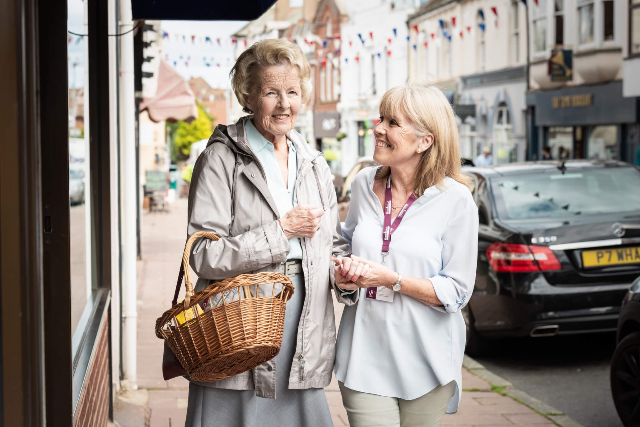 An elderly woman and a caregiver smiling and talking while walking on a street with shops and decorated flags. - Home Instead