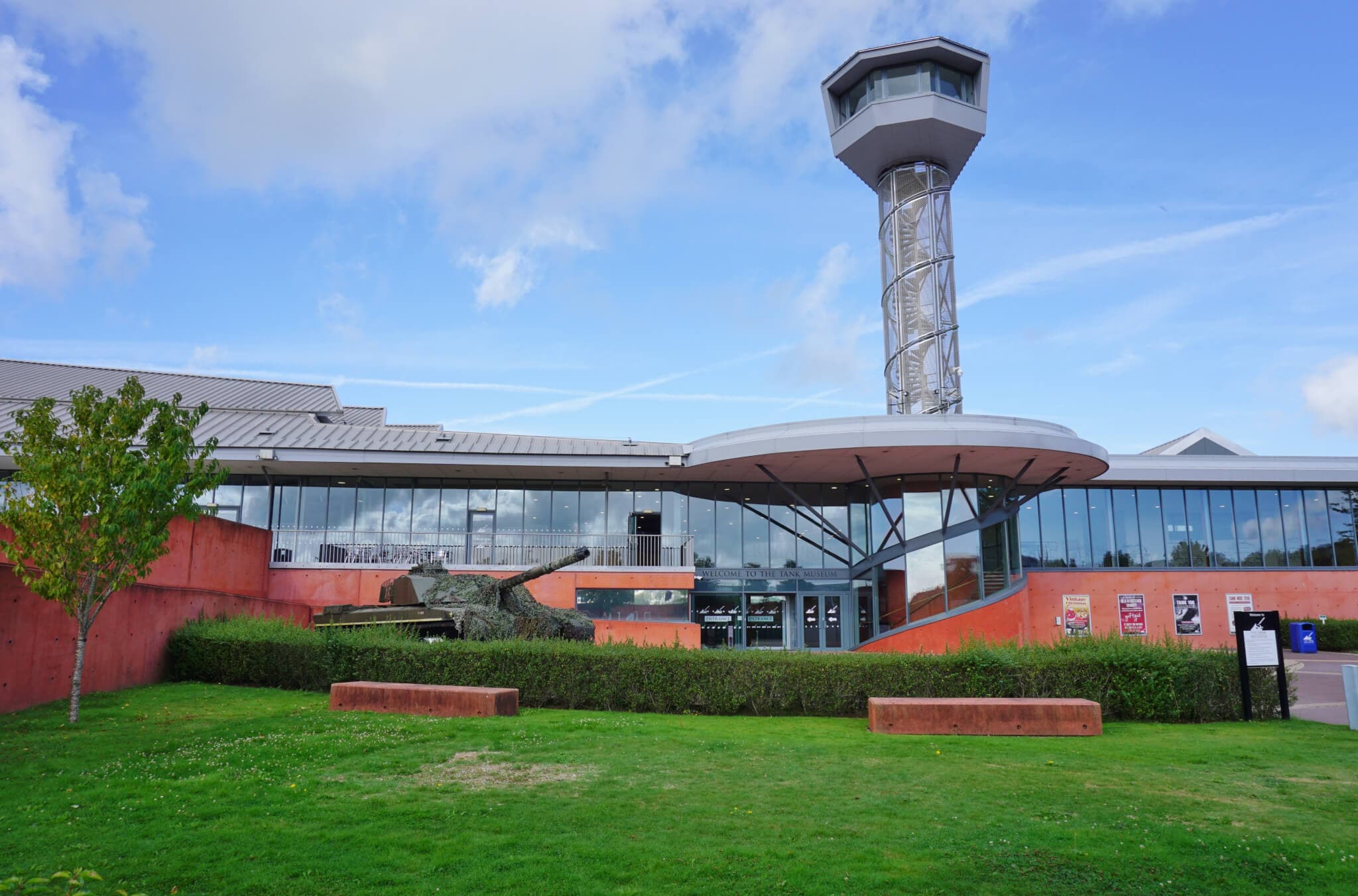 A modern building with a control tower, glass facade, and an outdoor display of a military tank on a grassy lawn. - Home Instead