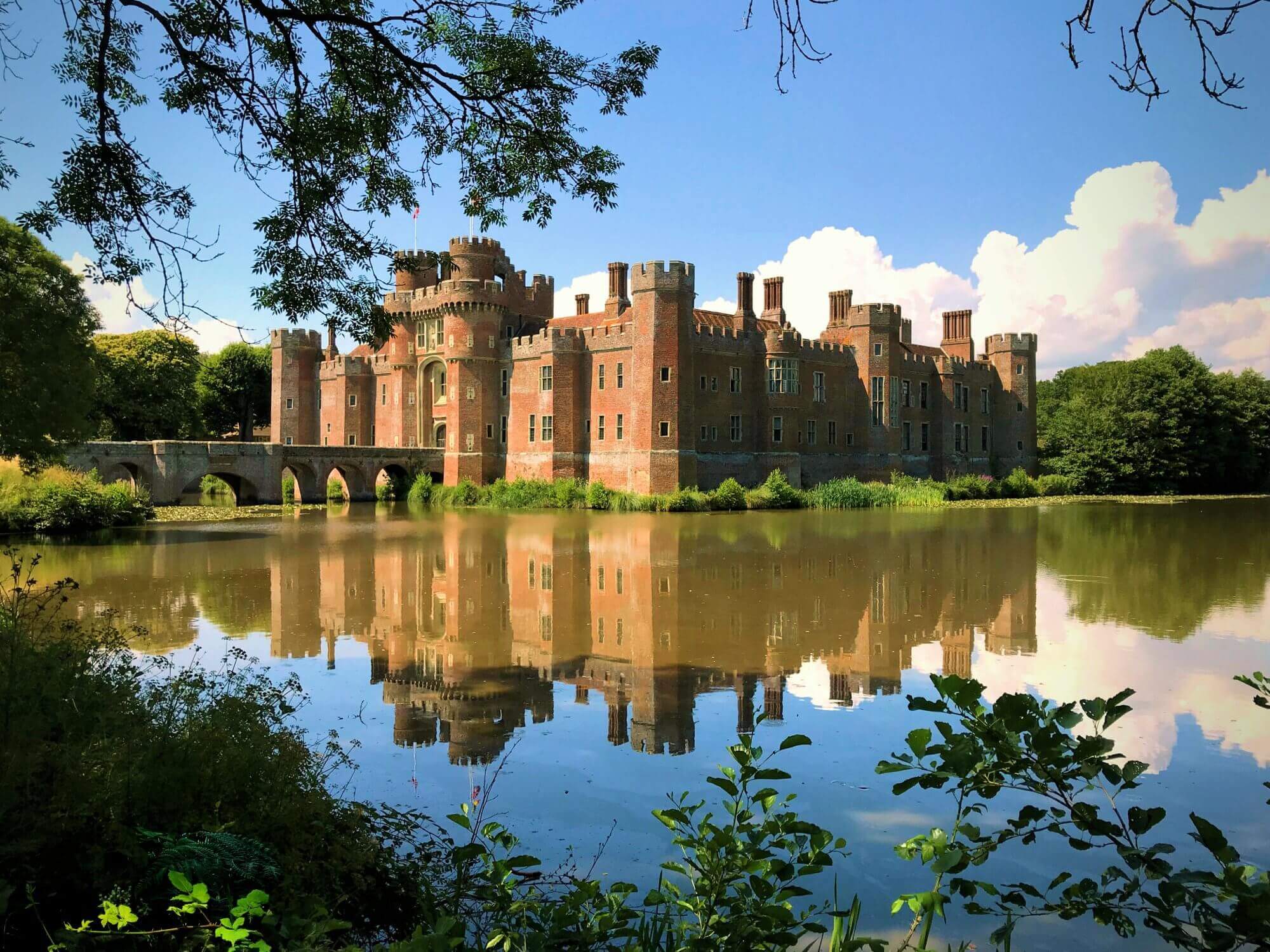 Brick castle with towers and battlements by a lake, its reflection visible in calm water, framed by trees and foliage. - Home Instead