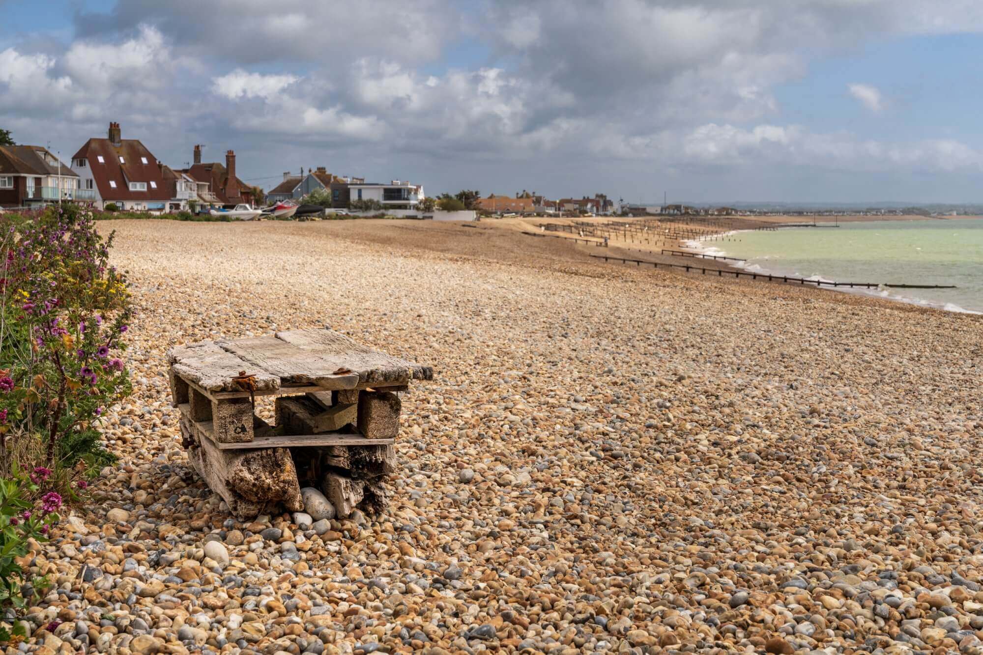 A pebble beach with houses in the background and a rustic wooden bench in the foreground on an overcast day. - Home Instead