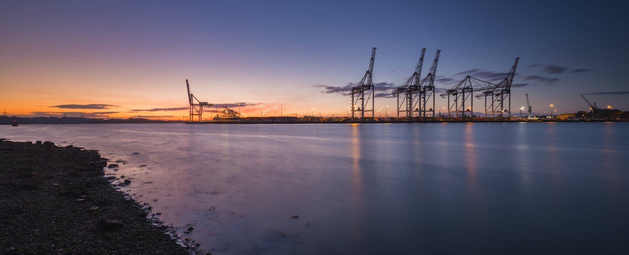 Sunset over a harbor with cranes silhouetted against the sky, and calm water reflecting the colorful evening light. - Home Instead