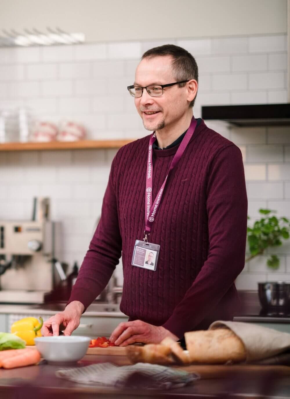 Man with glasses in a maroon sweater, wearing a lanyard, standing in a kitchen chopping vegetables. - Home Instead