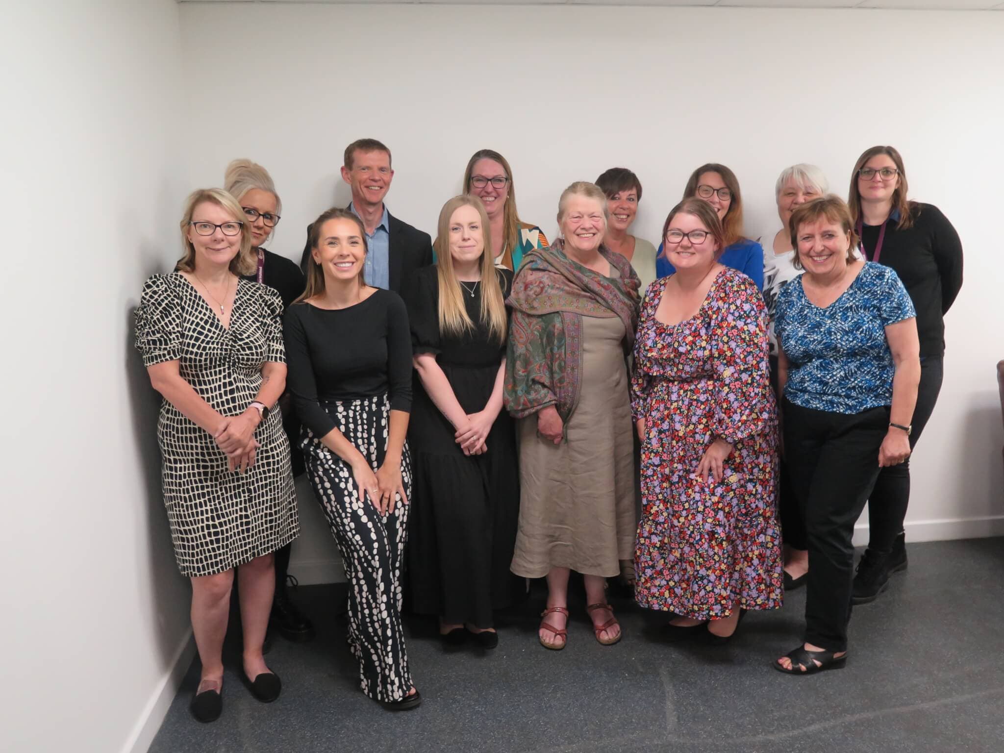 A group of 14 people, smiling, standing together in a brightly lit room with a plain white wall in the background. - Home Instead