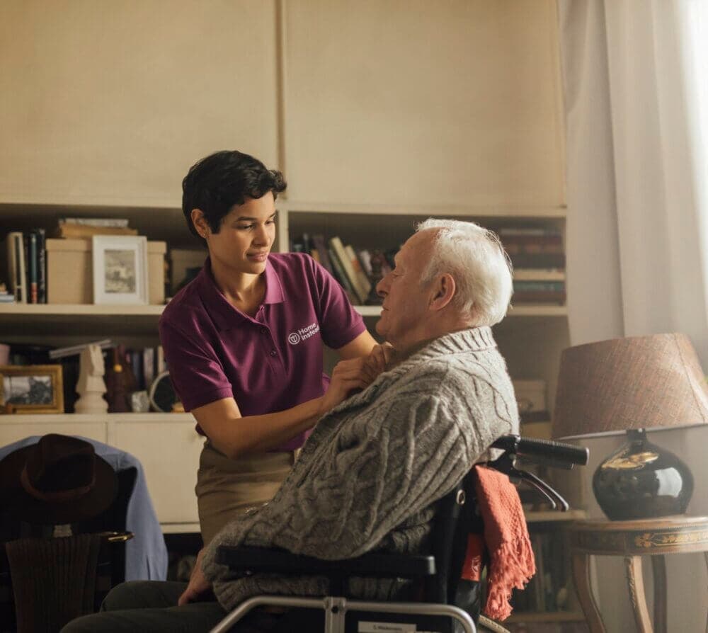 Caregiver in a purple shirt assisting an elderly man in a wheelchair with his sweater in a cozy living room. - Home Instead