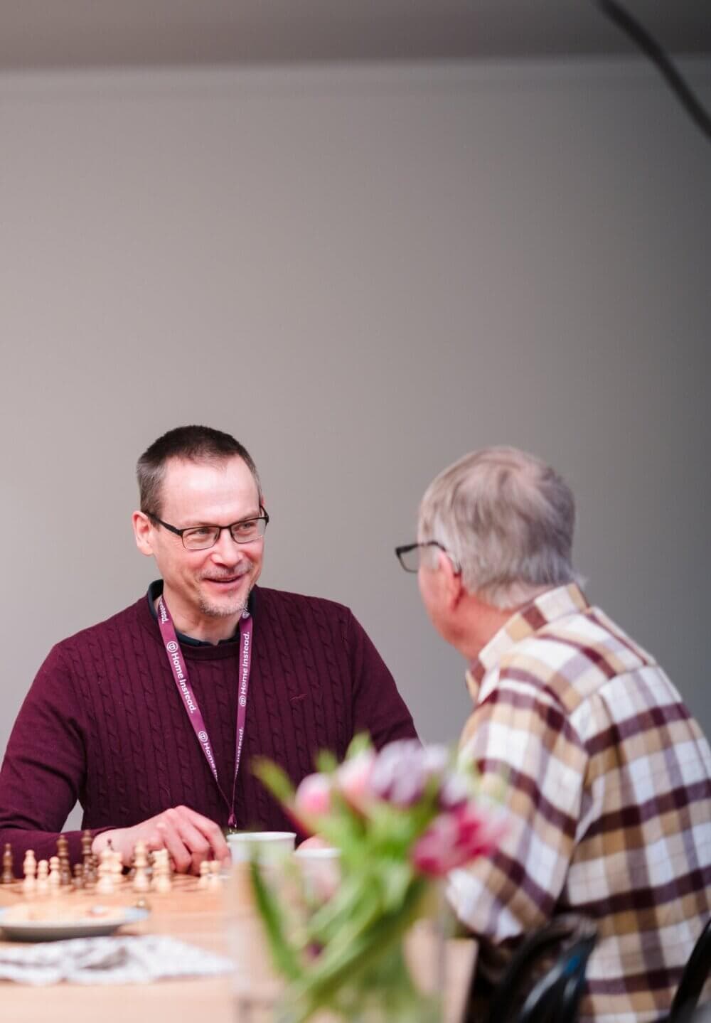 Two elderly men playing chess at a table with a bouquet of flowers in the foreground. - Home Instead