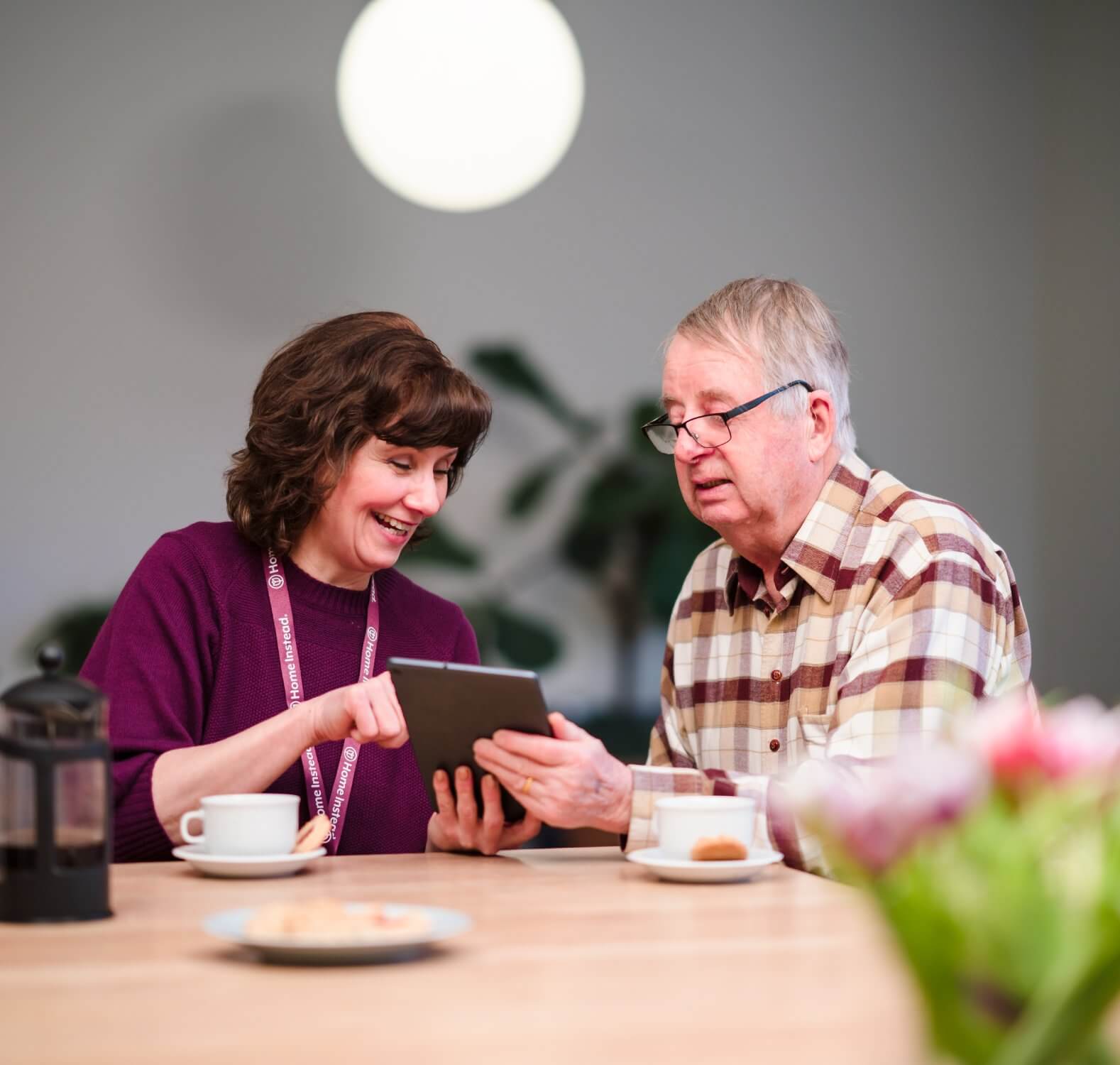 A woman showing an older man something on a tablet at a table with coffee and flowers. - Home Instead