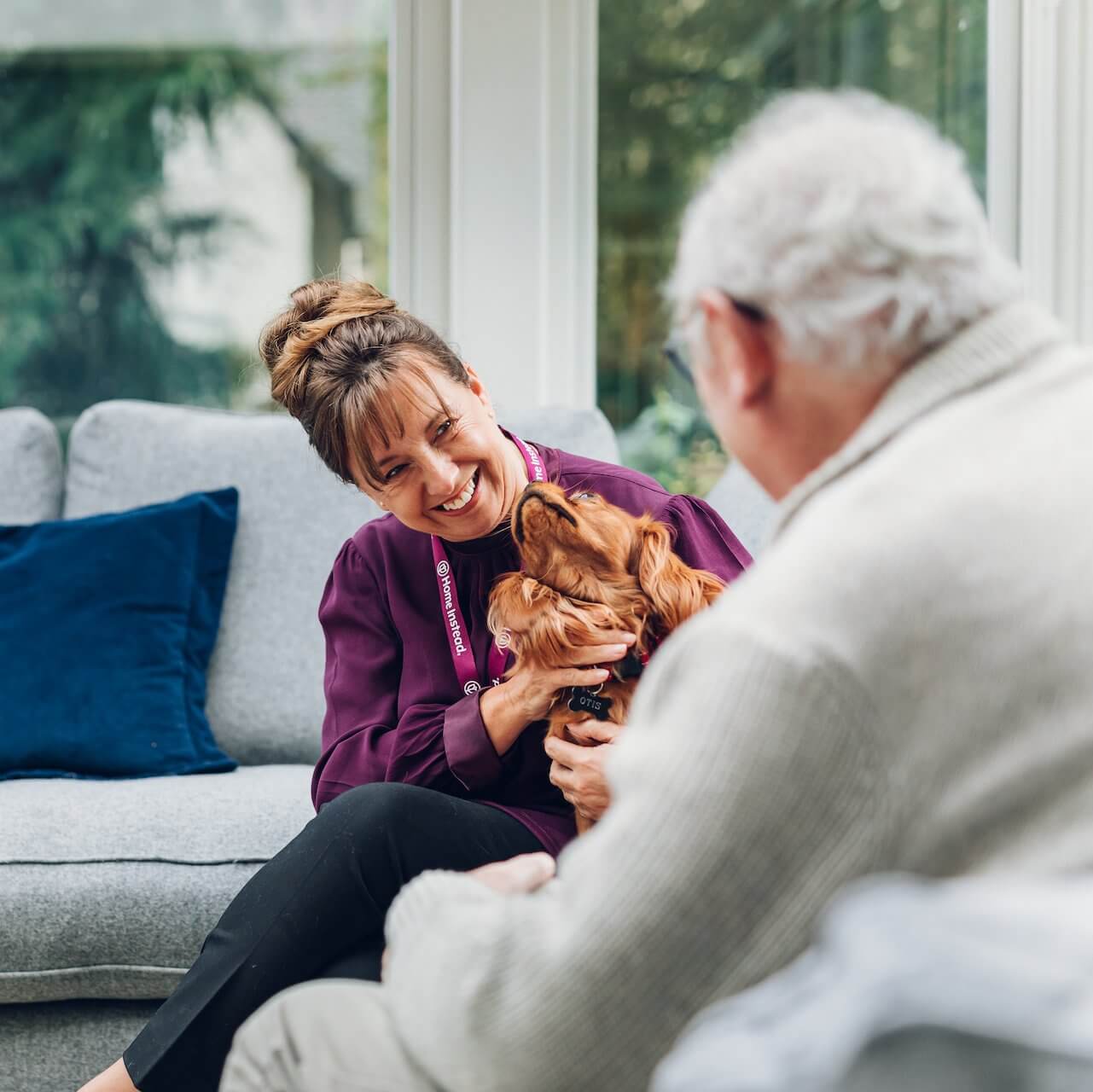 A woman and an older man sit on a couch, smiling at a dog in the woman's arms. - Home Instead