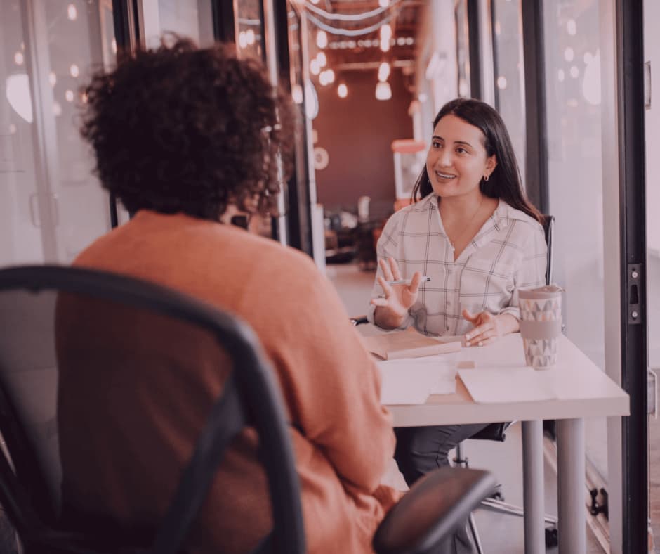 Two women sitting at a desk in an office, engaged in a conversation, with a mug and papers on the table. - Home Instead