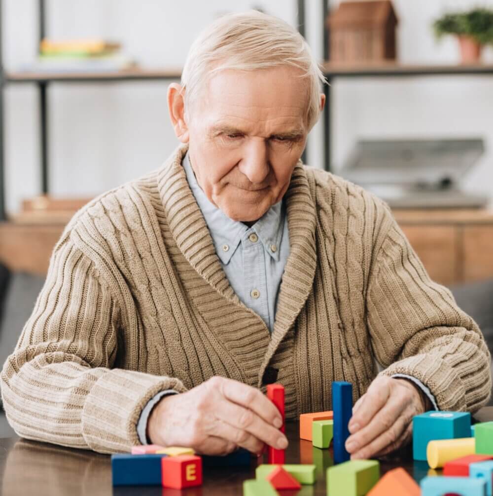 Elderly man in a beige sweater playing with colorful building blocks at a table. - Home Instead