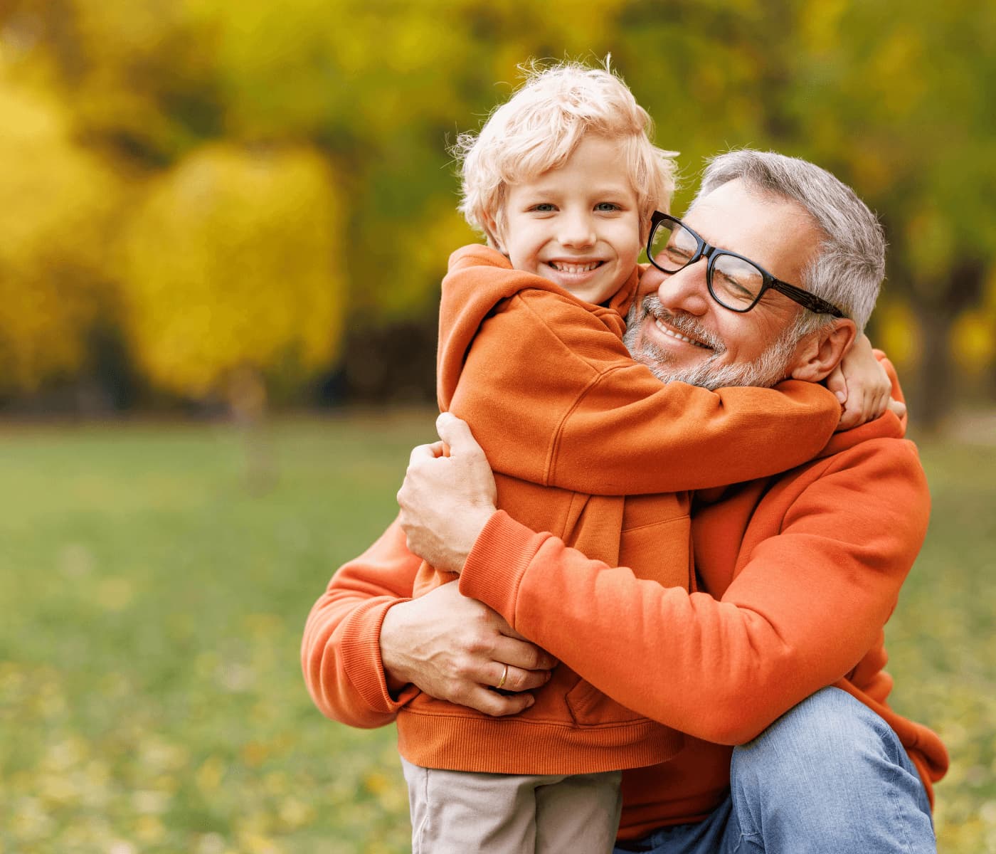 A smiling elderly man with glasses hugs a cheerful child outdoors in a park, both wearing orange hoodies. - Home Instead