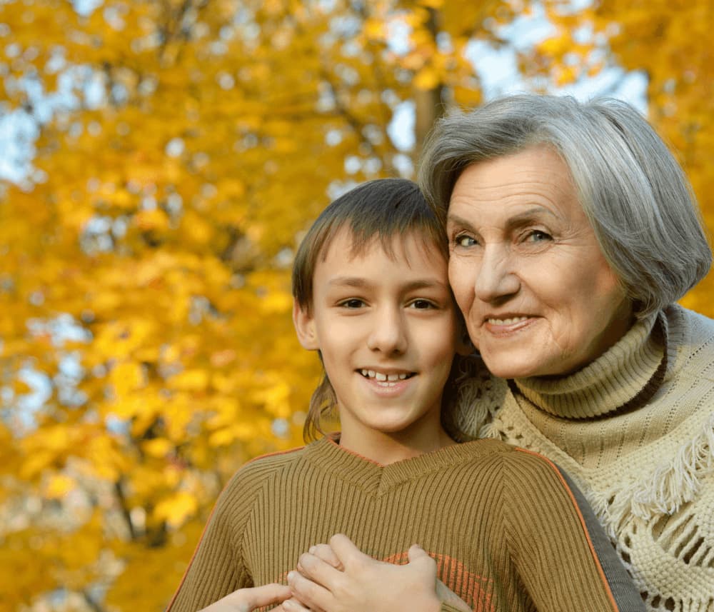 An elderly woman and a young boy smiling outdoors with autumn leaves in the background. - Home Instead
