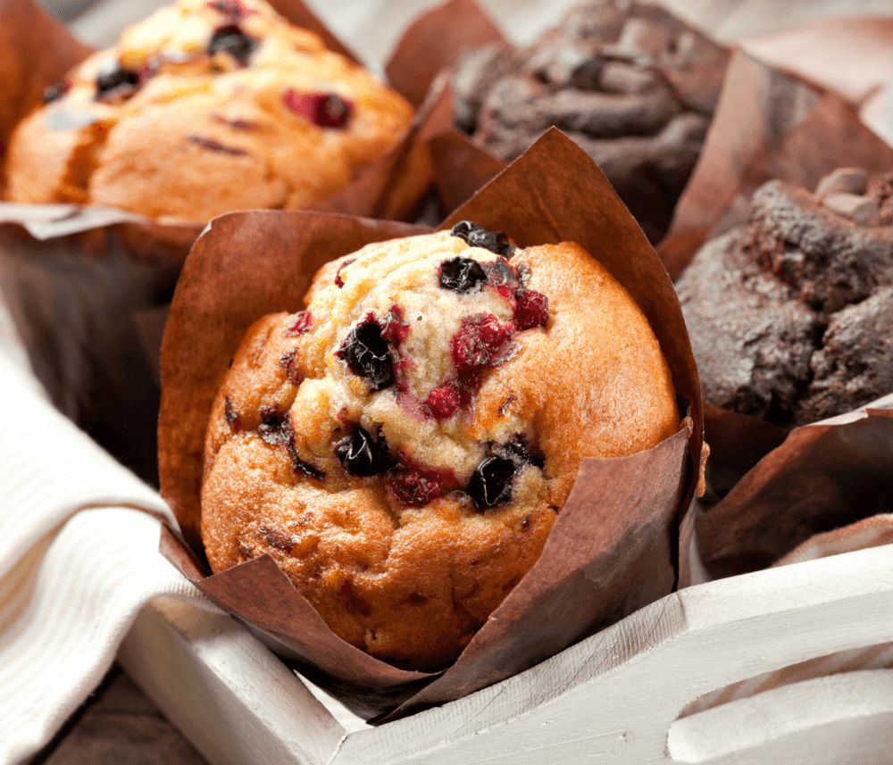 A close-up of assorted muffins in brown paper liners on a wooden tray, featuring berry and chocolate flavors. - Home Instead