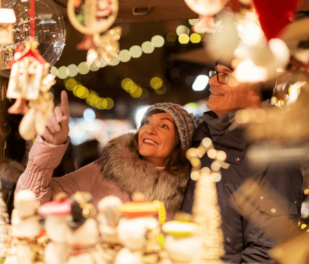 A couple enjoys browsing holiday decorations at a festive market stall, surrounded by bright, decorative ornaments. - Home Instead