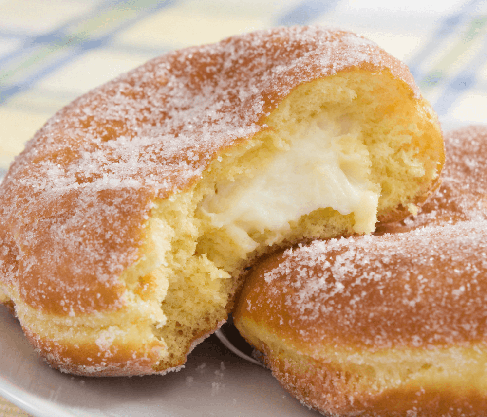 A sugar-coated donut filled with cream, partially bitten to reveal the creamy filling inside, on a white plate. - Home Instead