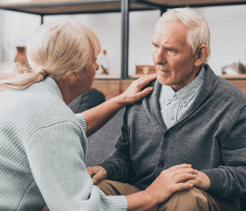Elderly woman comforting an elderly man by placing her hand on his shoulder and holding his hand. - Home Instead