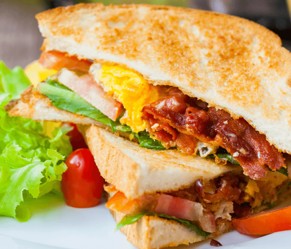 A BLT sandwich with crispy bacon, lettuce, tomato, and fried egg, served with a side salad on a white plate. - Home Instead