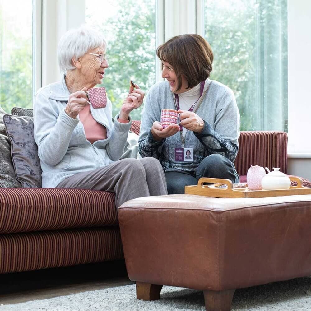 An elderly woman and a younger woman sit on a sofa, sharing a laugh while drinking tea in a sunlit room. - Home Instead