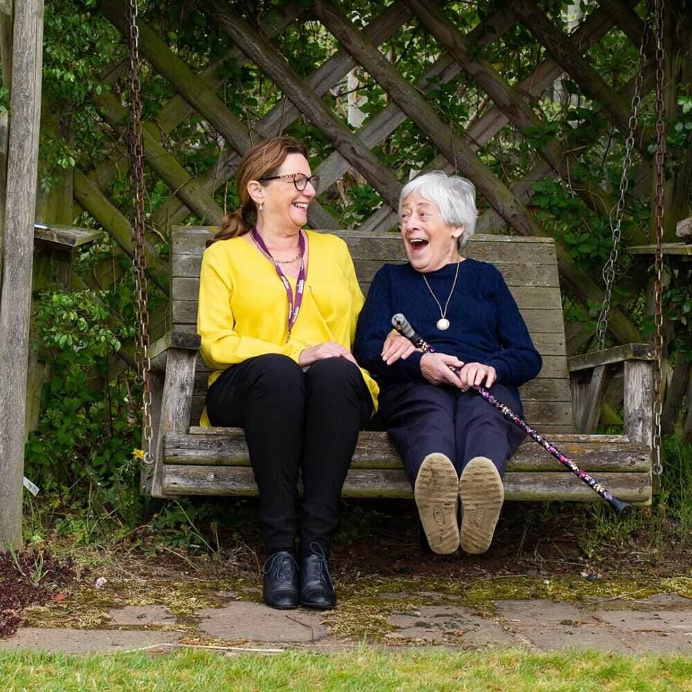Two women sitting on a wooden swing, laughing and enjoying a moment outdoors near a trellis with greenery. - Home Instead