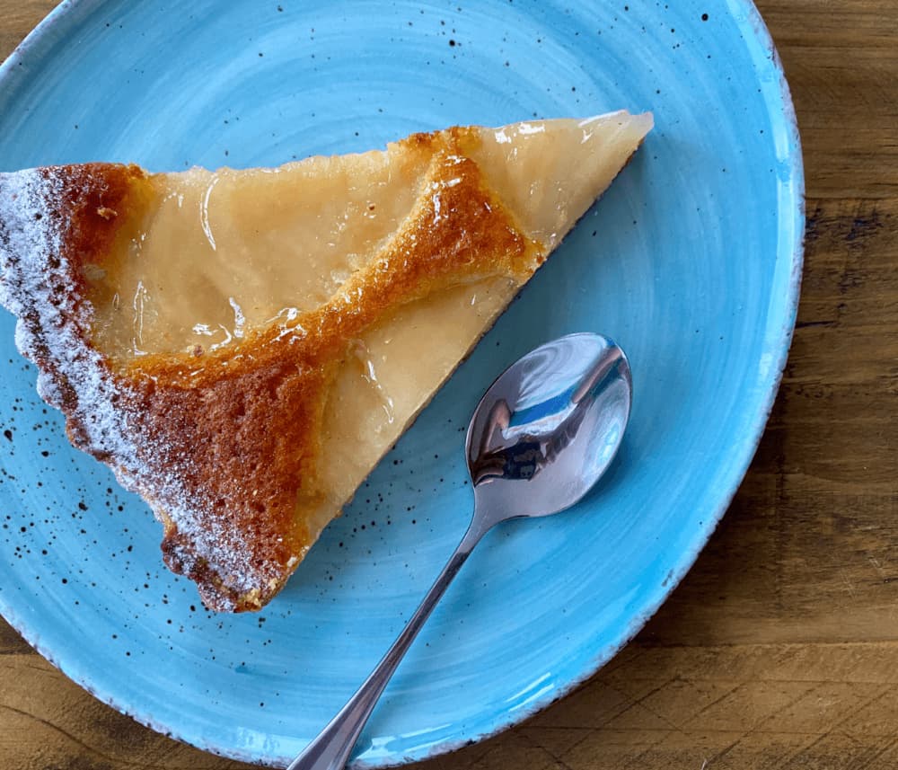 A slice of pear tart on a blue plate with a teaspoon beside it, placed on a wooden table. - Home Instead