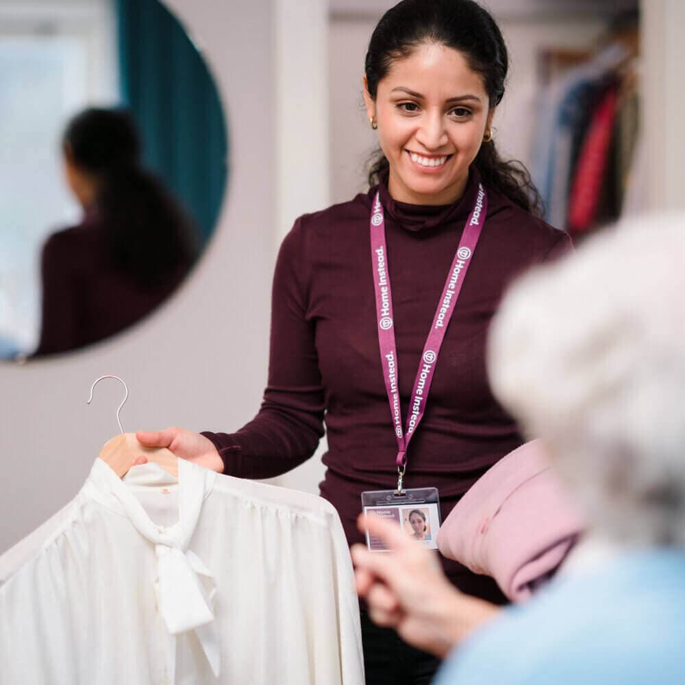 A smiling woman holding a white blouse on a hanger and assisting another person, who is partially visible. - Home Instead