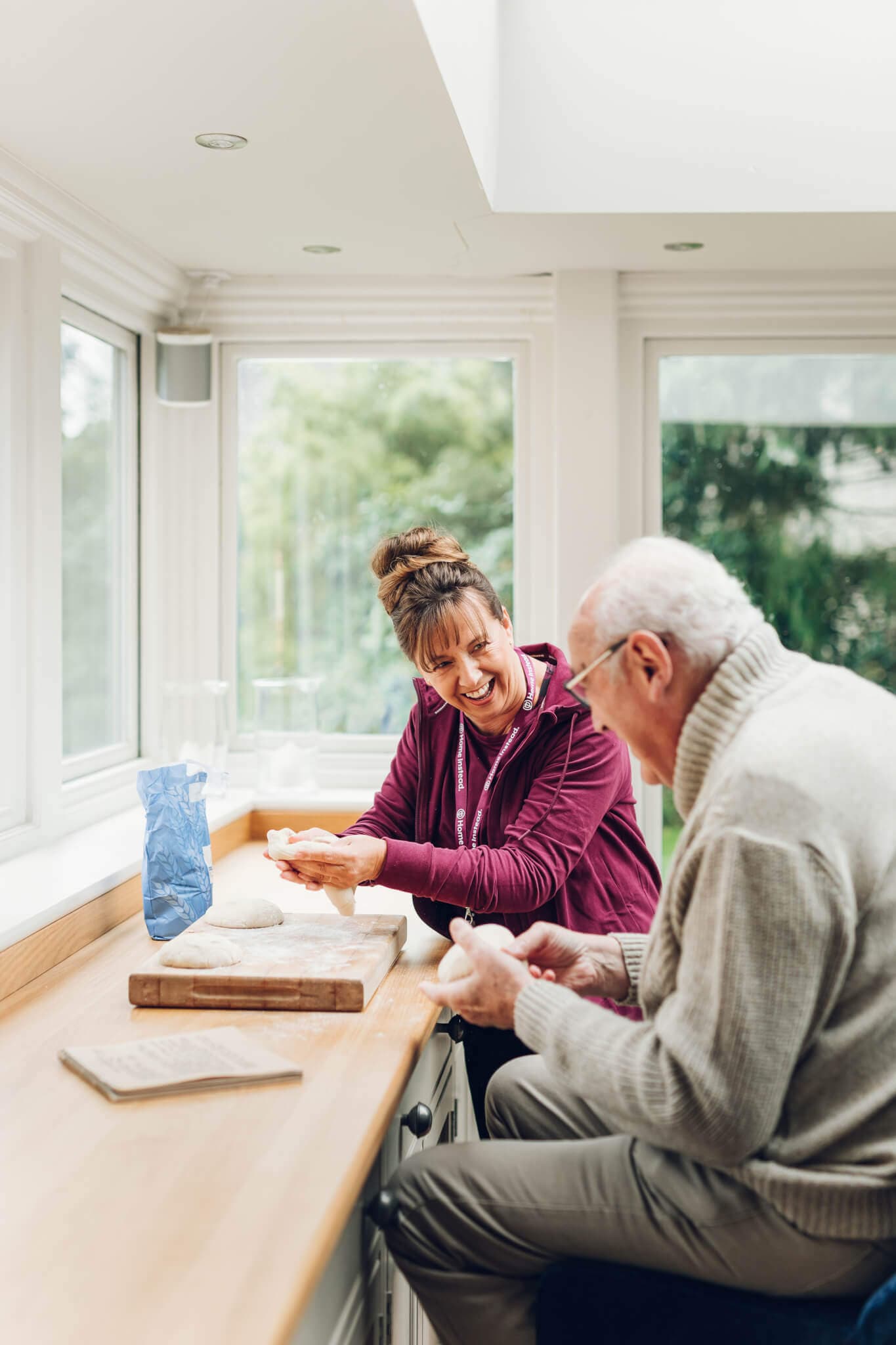 A caregiver and elderly man smile as they bake together in a bright kitchen with large windows. - Home Instead