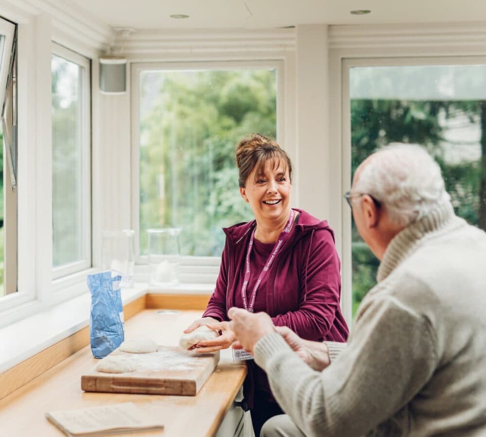 Woman and elderly man smiling and talking at a table in a bright room with large windows. - Home Instead Southampton