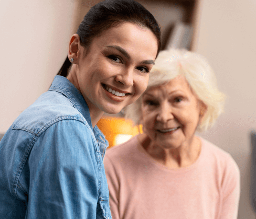 Young woman smiling with an elderly woman in the background, both looking at the camera, in a cozy indoor setting. - Home Instead