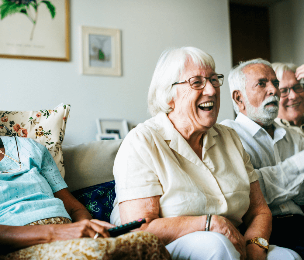 Four elderly people sitting on a couch, smiling and enjoying a moment together in a living room setting. - Home Instead
