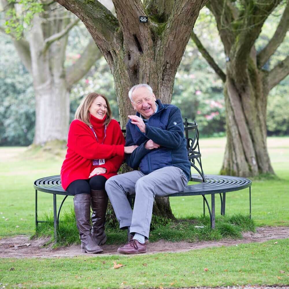Two people, smiling and seated on a circular bench around a large tree in a park, share a joyful moment. - Home Instead