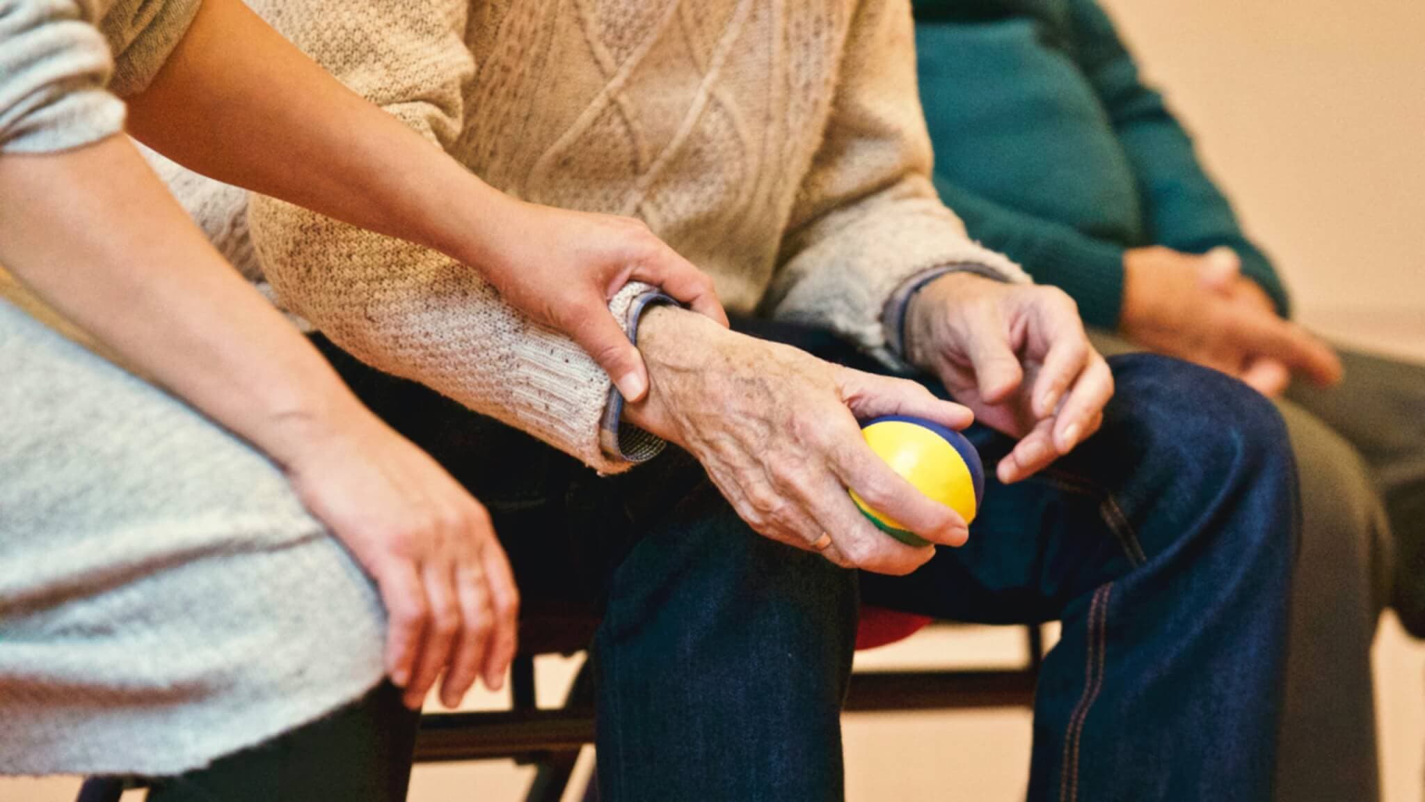 A person helps an elderly individual holding a ball, providing support by gently holding their arm. - Home Instead