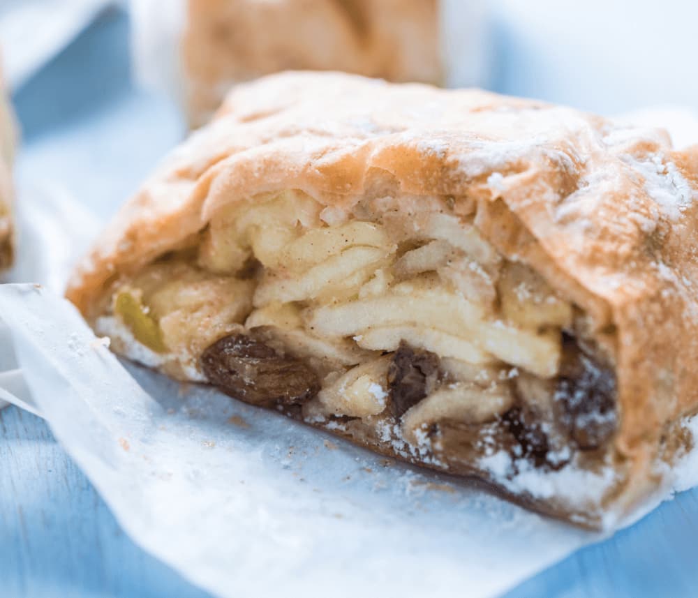 Close-up of a slice of apple strudel with visible layers of apples and raisins within flaky pastry, dusted with powdered sugar. - Home Instead