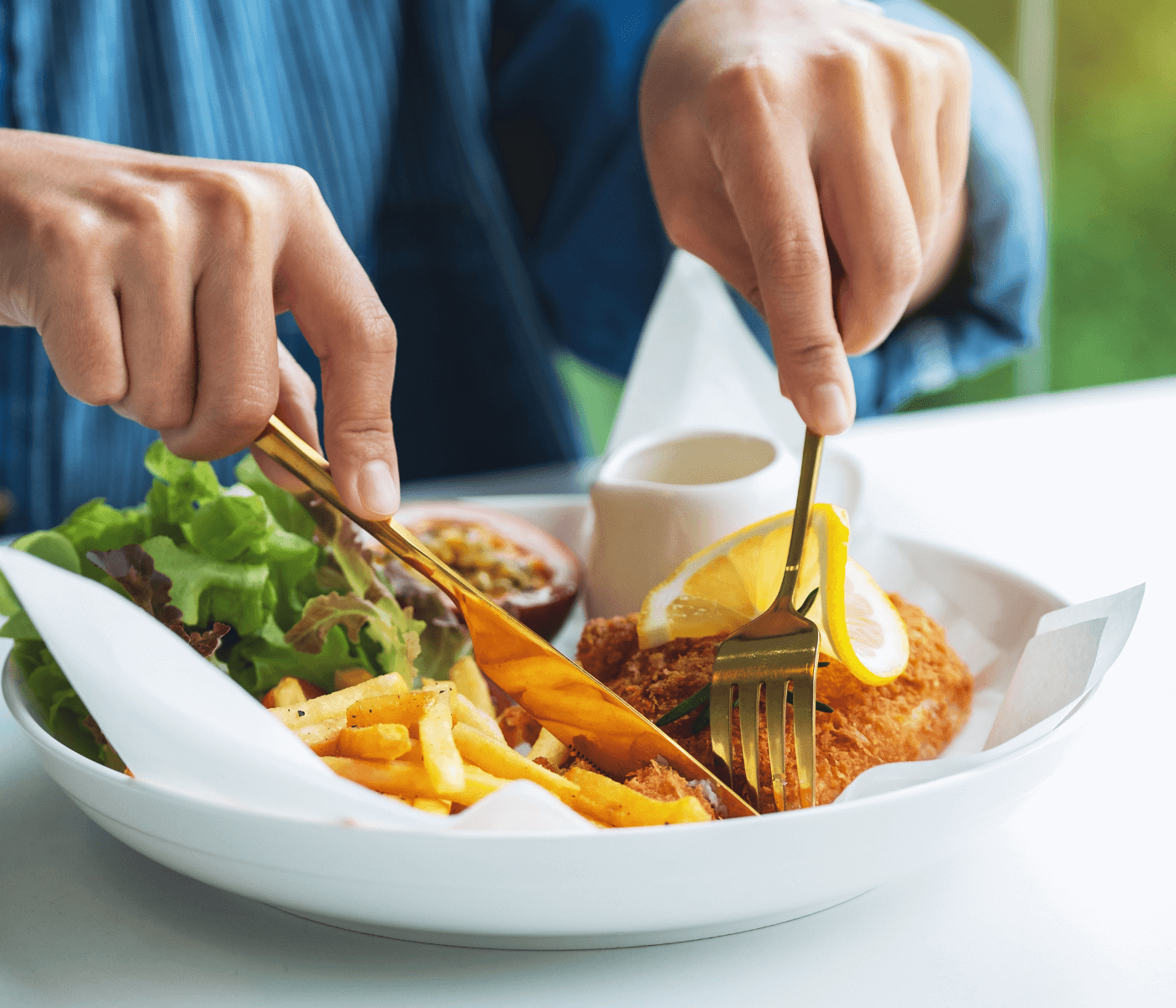 A person cuts into breaded fish in a bowl with fries, greens, and a lemon slice on a table, using gold utensils. - Home Instead