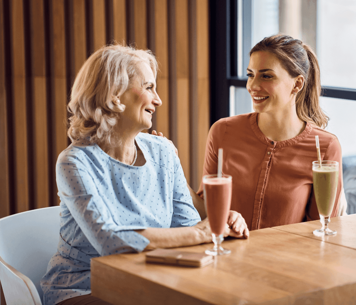 Two women sitting at a table, smiling and drinking smoothies, with one woman resting a hand on the other's shoulder. - Home Instead