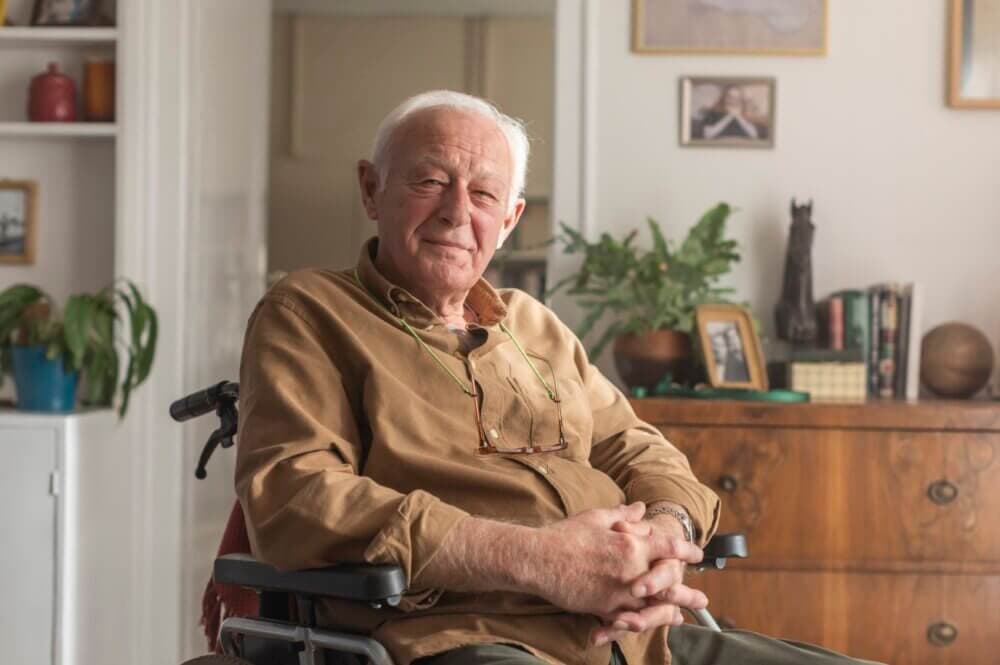 Elderly man in a brown shirt sitting in a wheelchair, smiling in a cozy, plant-decorated room. - Home Instead