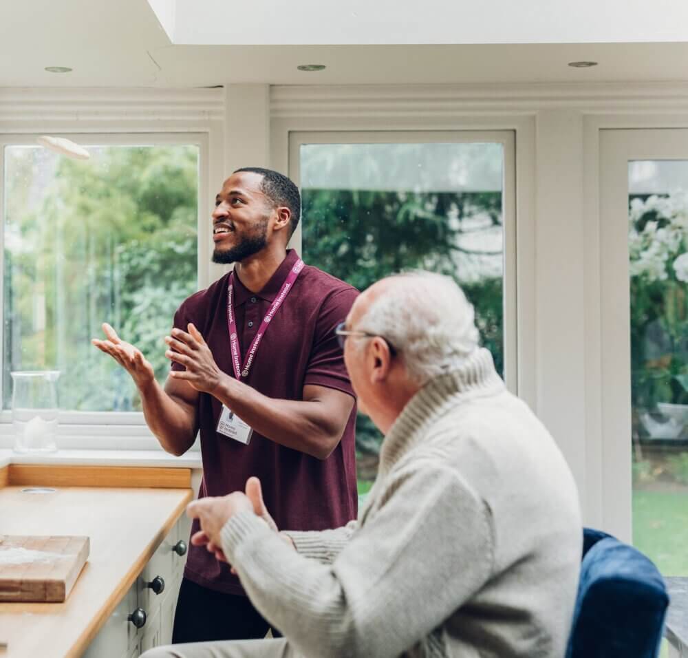 A caregiver and an elderly man are in a bright room; the caregiver is smiling and gesturing animatedly. - Home Instead