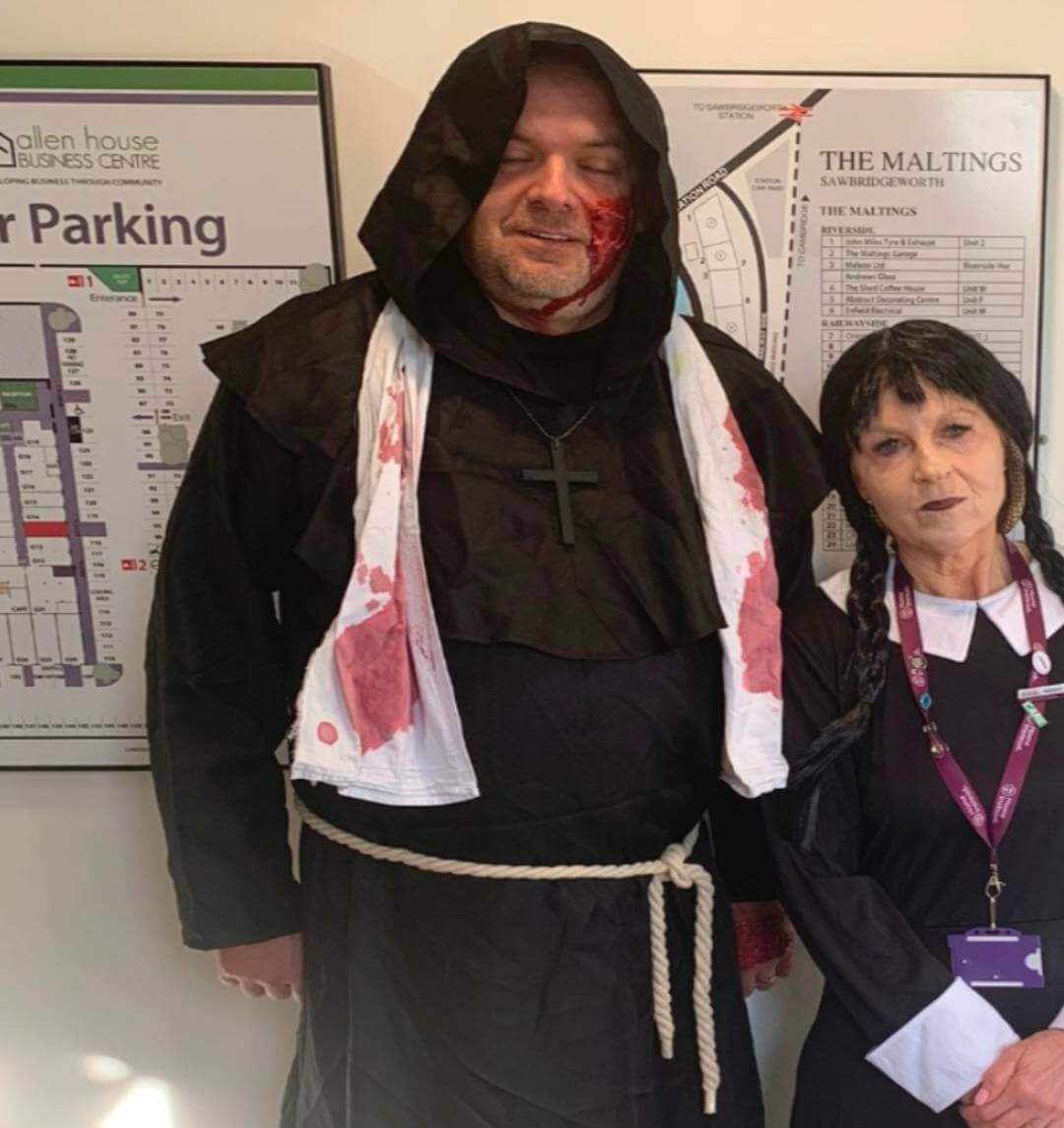 Two people in costumes: a man in a monk outfit with a bloody face and a woman in a black dress with braids. - Home Instead