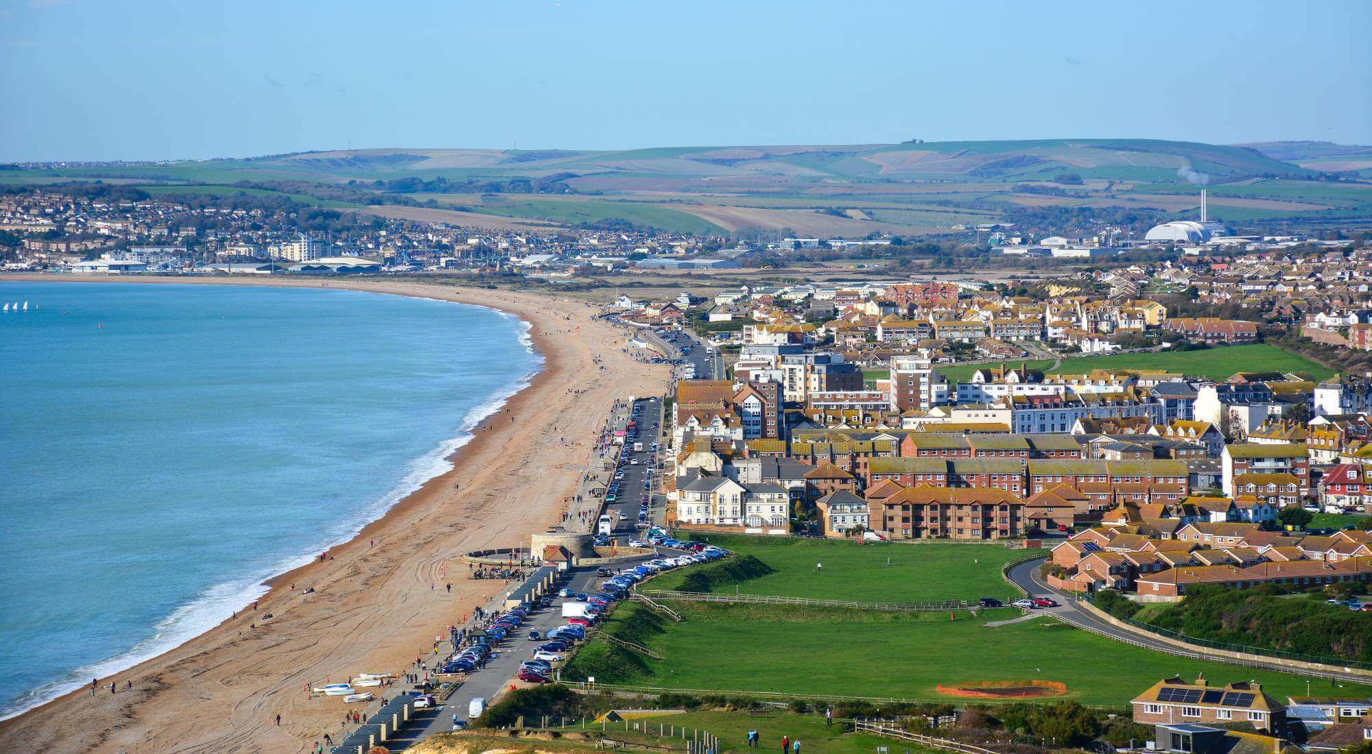 Aerial view of a coastal town with a beach, green hills, and various buildings along the shoreline. - Home Instead