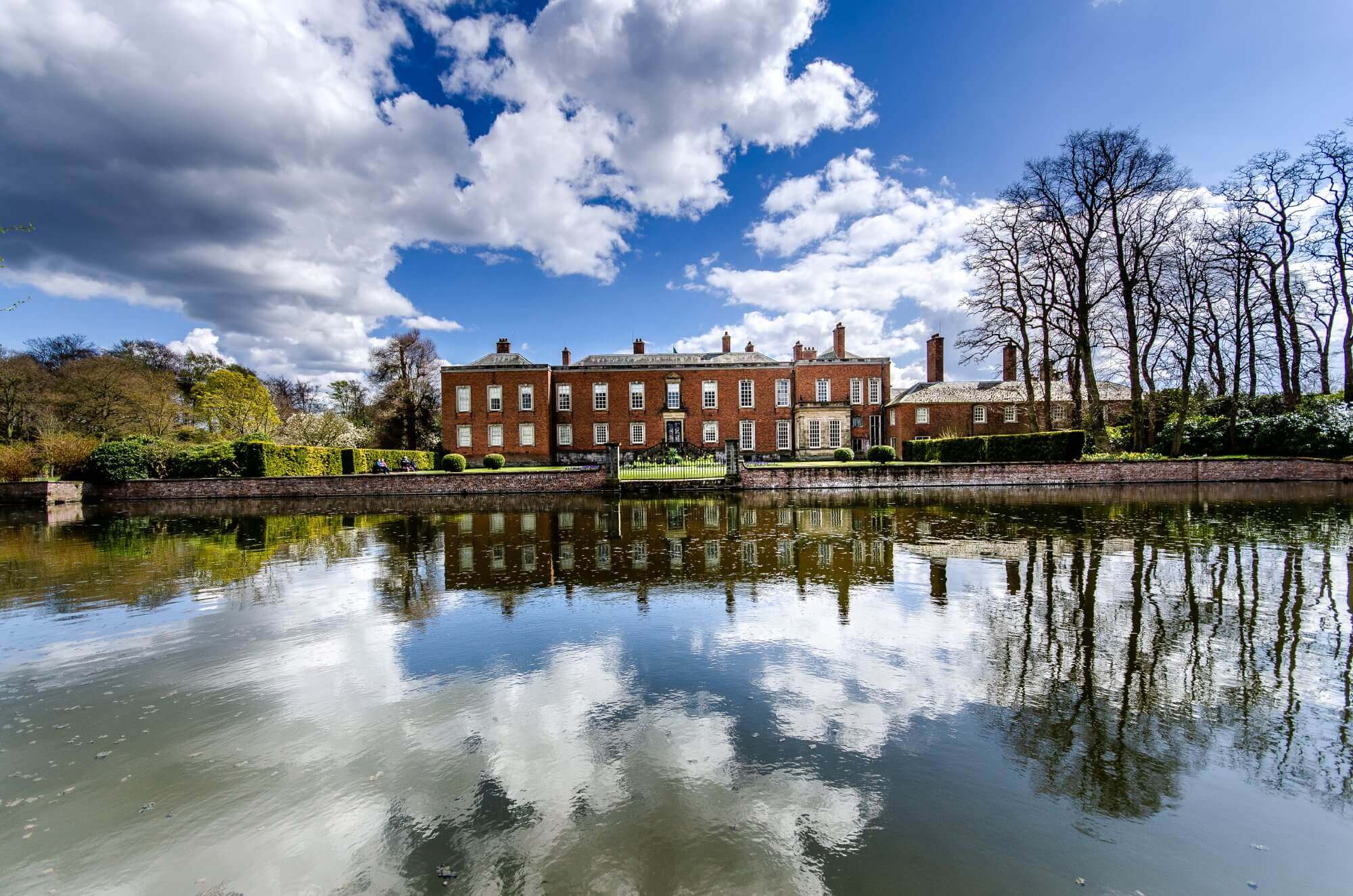 Grand brick mansion with multiple chimneys, reflected in a large, still pond, under a sky filled with dramatic clouds. - Home Instead
