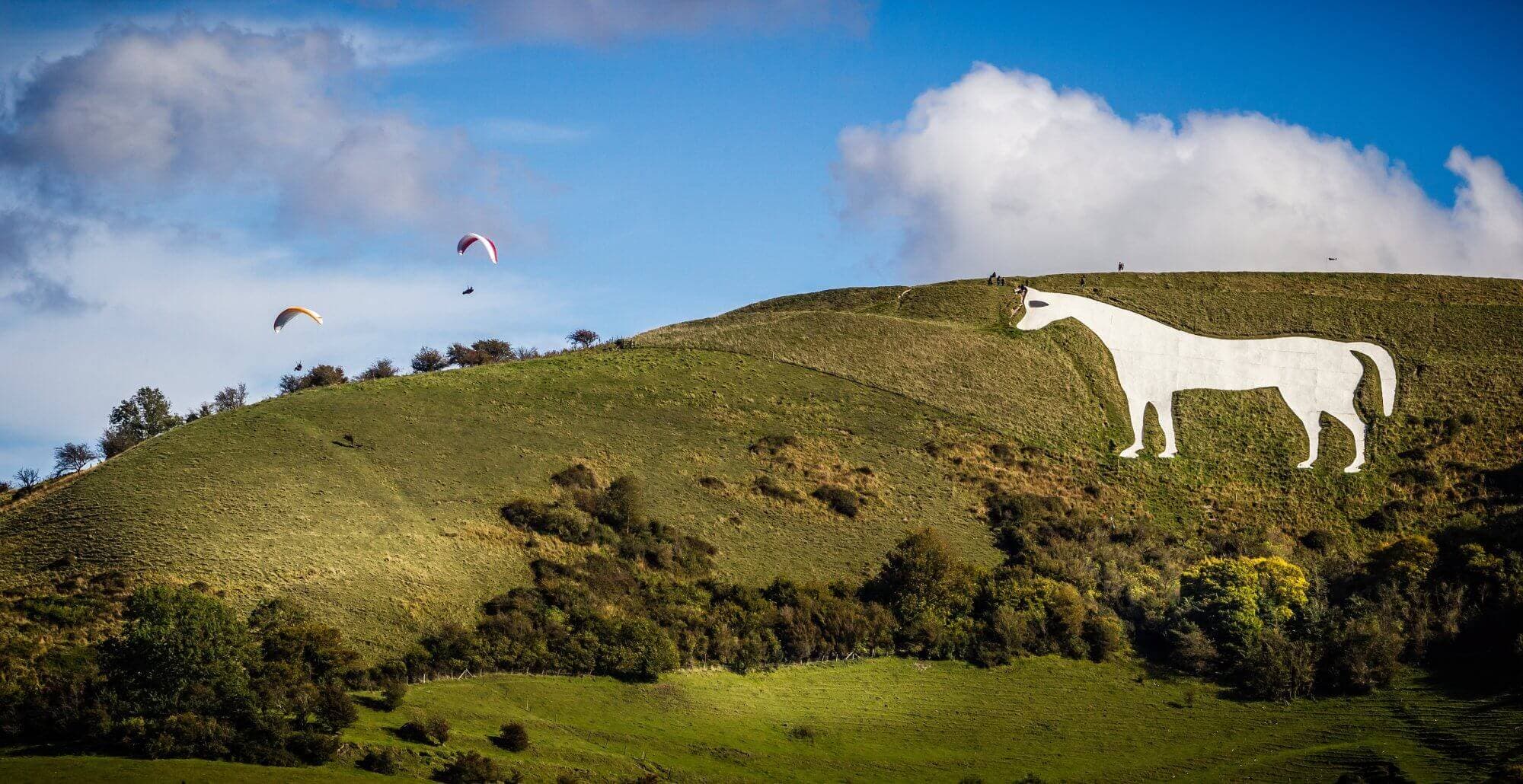 White horse hill figure on a green hillside with three paragliders floating in the sky above. - Home Instead