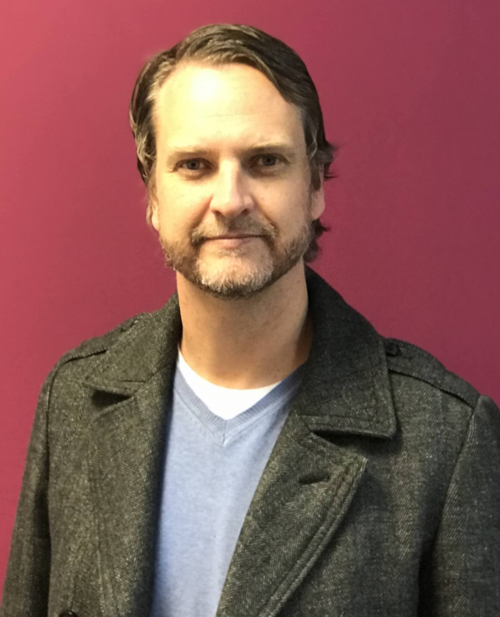 Man with short hair and a beard wearing a gray coat and a blue shirt stands in front of a purple wall. - Home Instead