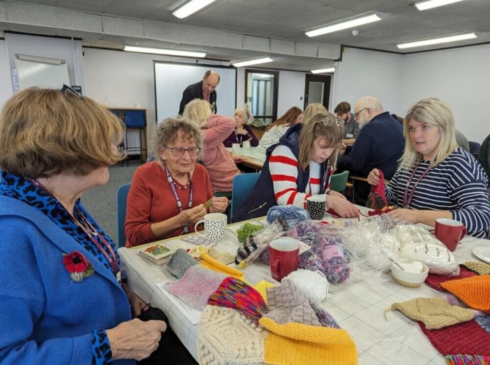 A group of people sitting around a table, knitting and chatting in a communal room. - Home Instead