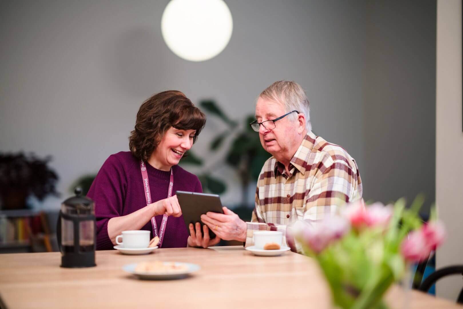 A woman helps an elderly man with a tablet at a table with coffee and flowers. - Home Instead