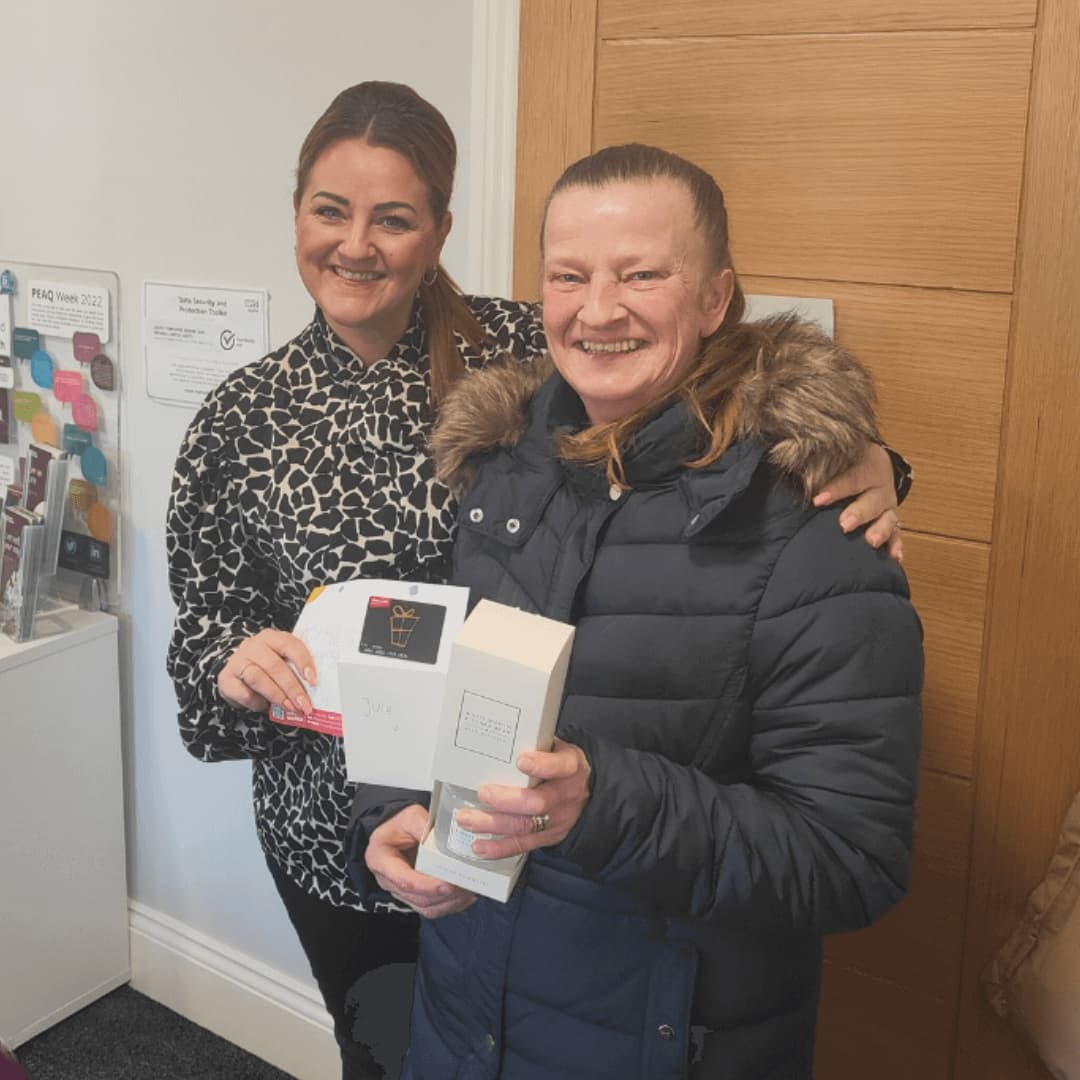 Two women smiling, one holding a small box and card, standing indoors in a casual setting. - Home Instead