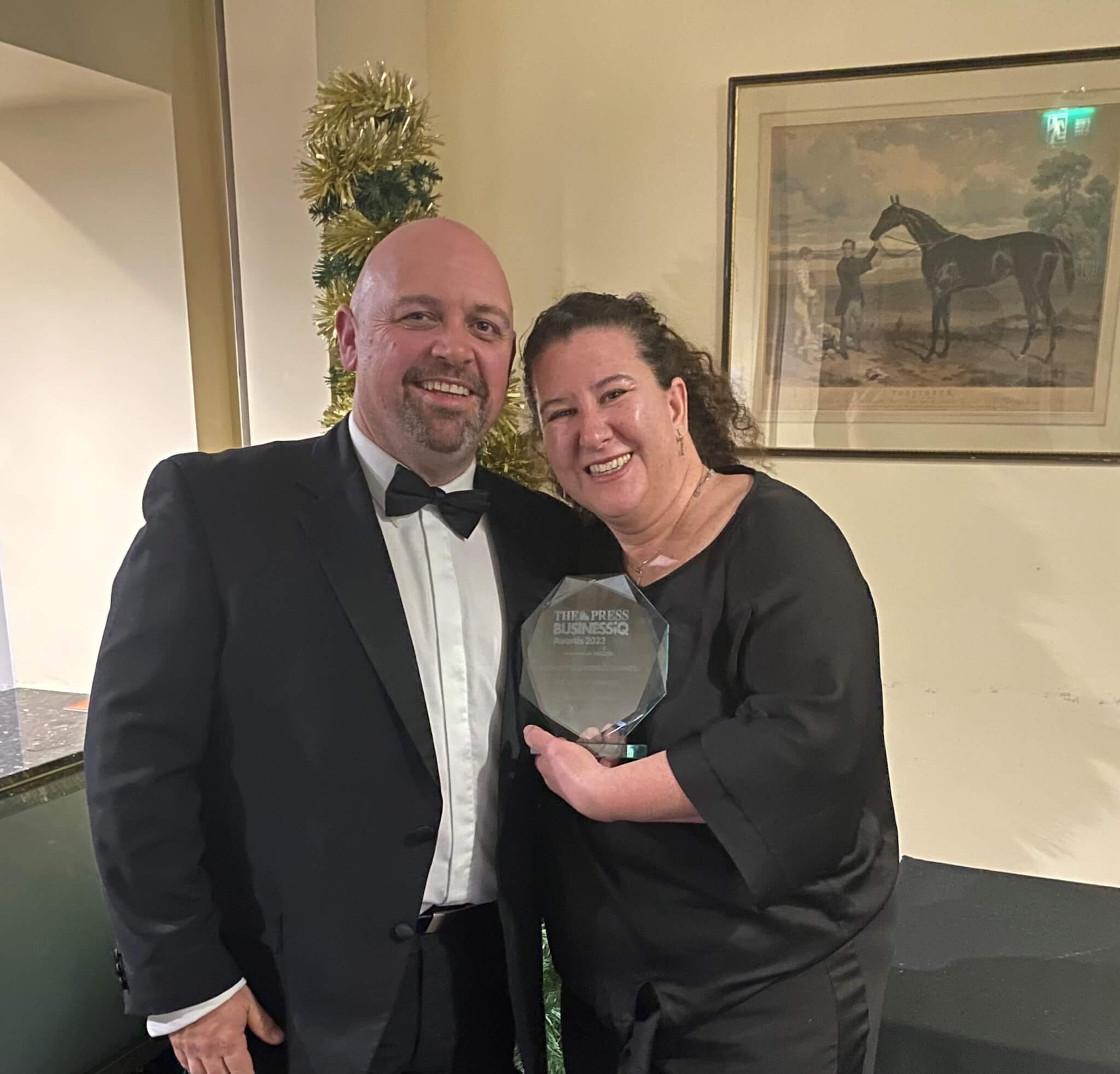 Two people dressed in formal attire smile, while one holds an award. A framed horse painting is on the wall behind them. - Home Instead