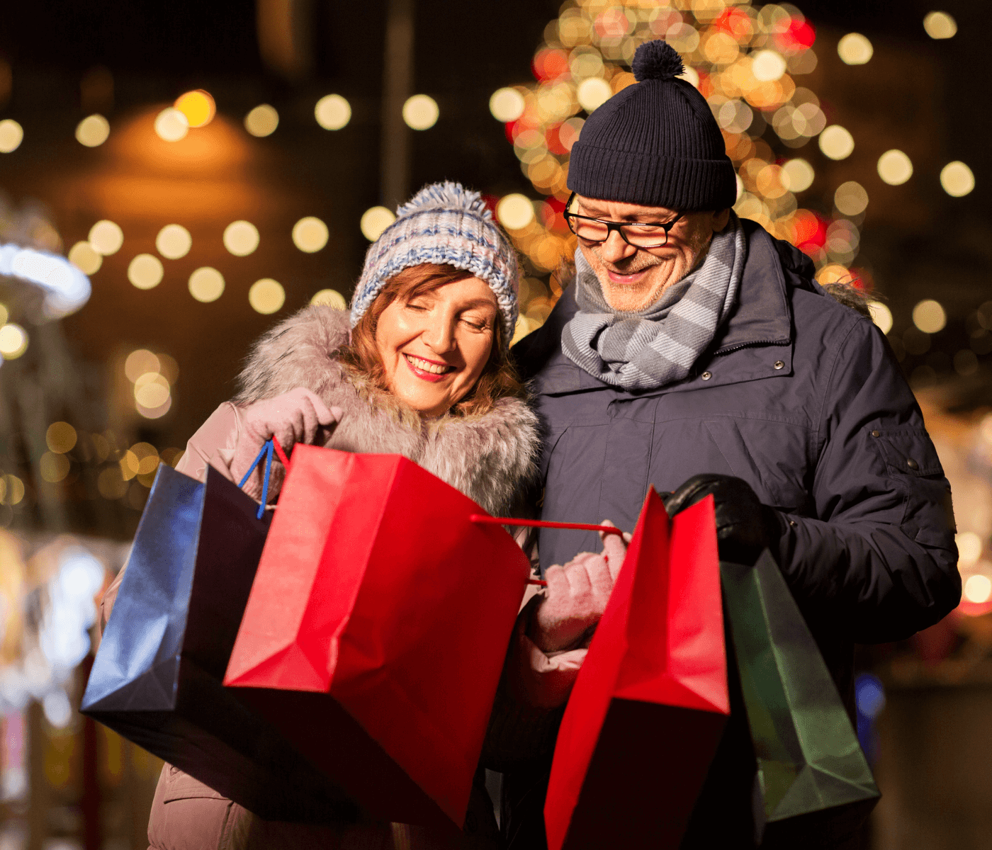 A smiling couple in winter clothes holds shopping bags and looks at them, with festive lights in the background. - Home Instead