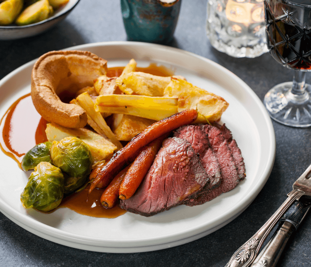 A plate of roast beef, Brussels sprouts, carrots, roasted potatoes, and Yorkshire pudding with a glass of red wine. - Home Instead