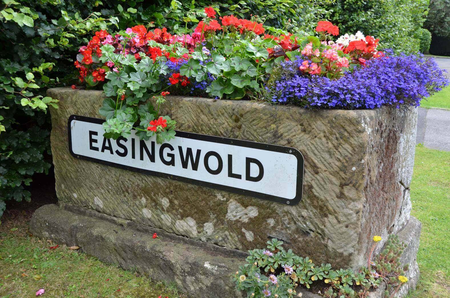 A stone planter with colorful flowers and a sign that reads "Easingwold" in a garden setting. - Home Instead