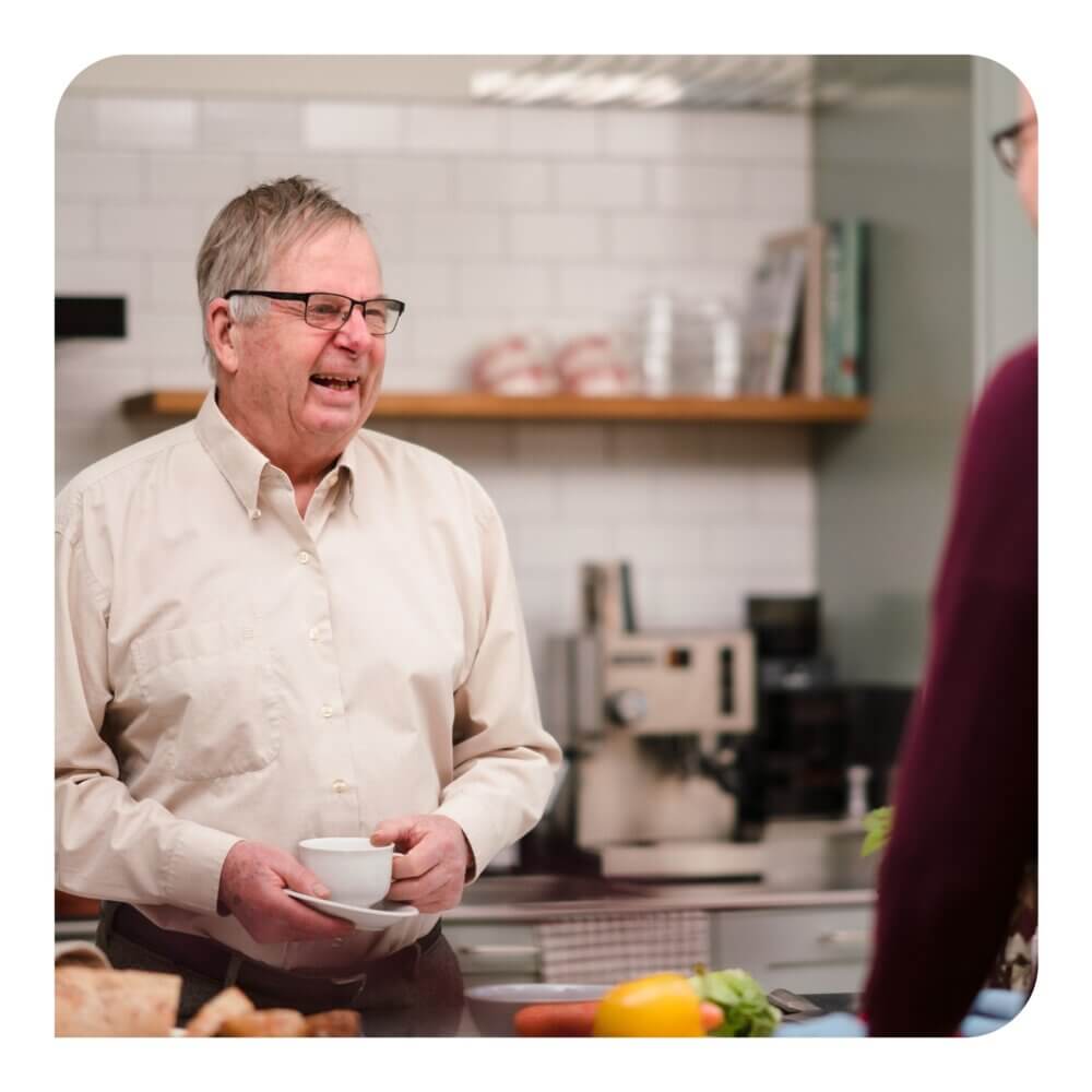 An elderly man in a kitchen holds a white cup, smiling and conversing with someone off-camera. - Home Instead