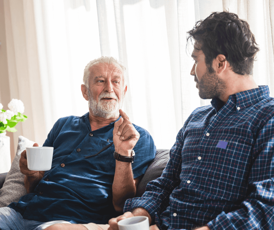 Older man and younger man sitting on a couch, talking and holding white mugs. - Home Instead