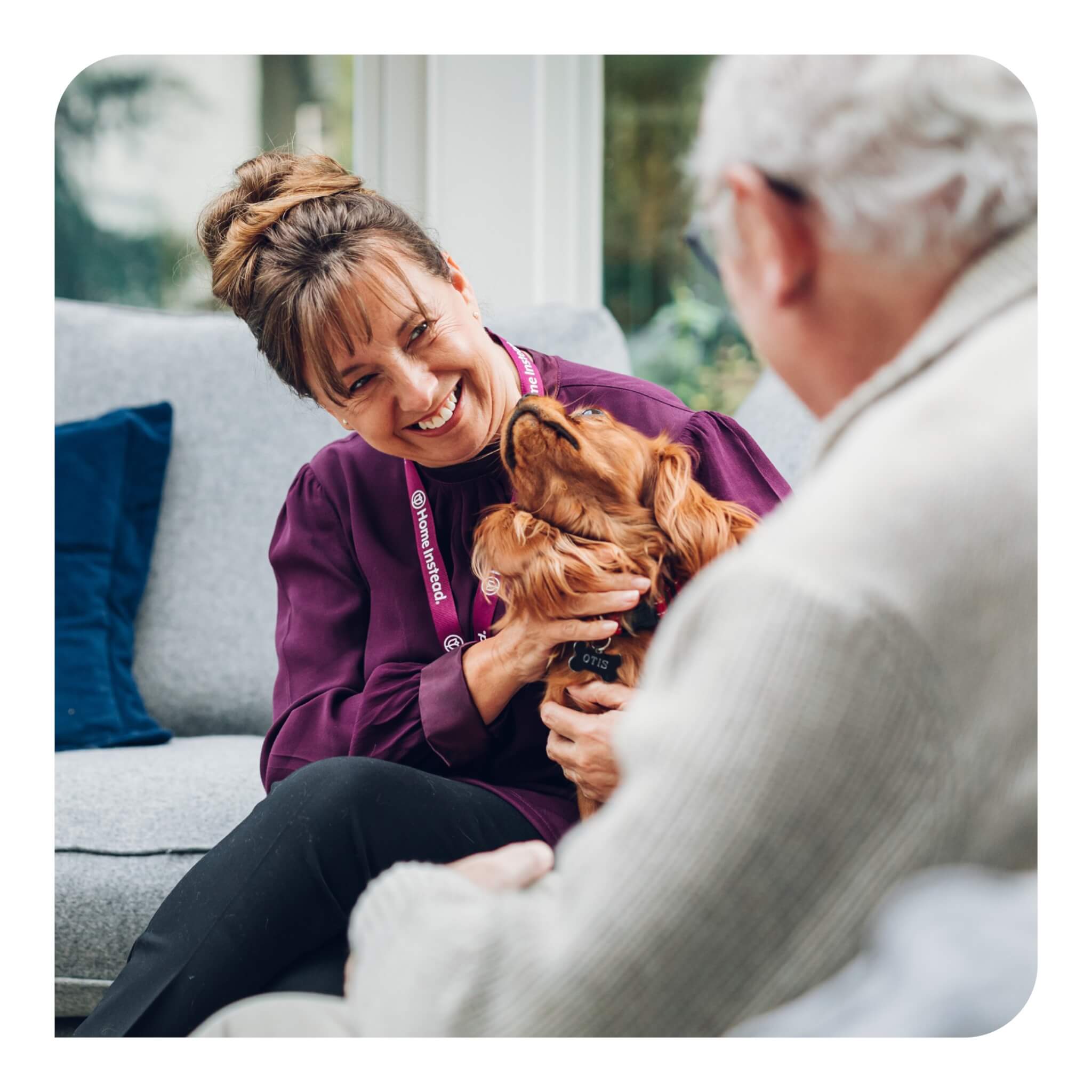 A woman with a name badge smiles while holding a dog and talking to an elderly man sitting on a couch. - Home Instead